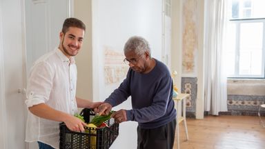 A young volunteer offers support by delivering fresh vegetables to an elderly gentleman indoors.