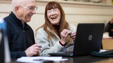 Joyful elderly couple sharing a laugh while using a laptop at a desk indoors.